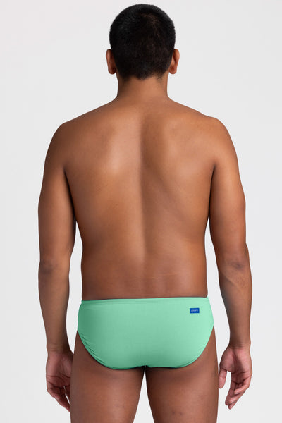Male model wearing the Men's BROLYN Brief Swimsuit in the Keylime solid, standing in front of a grey background, shown from back view. 
