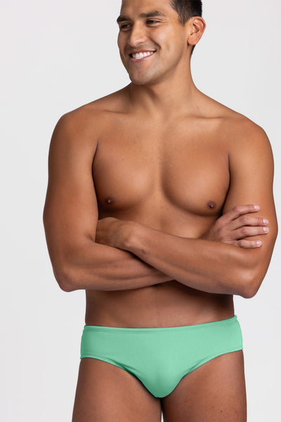Male model wearing the Men's BROLYN Brief Swimsuit in the Keylime solid, standing in front of a grey background, shown from front view. 
