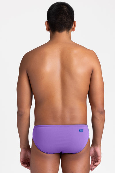 Male model wearing the Men's BROLYN Brief Swimsuit in the Paddle solid, standing in front of a grey background, shown from back view. 

