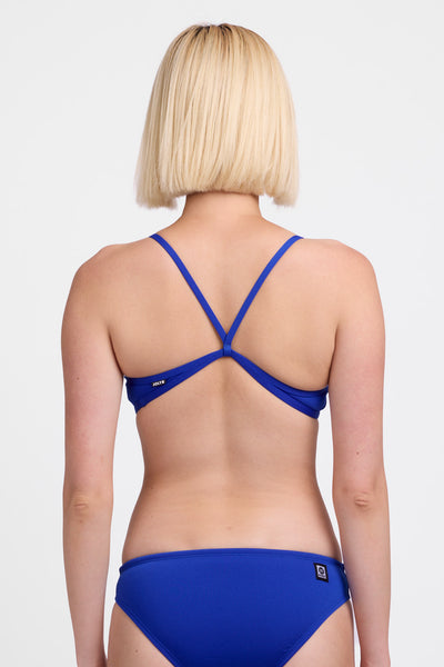 Female model wearing the Women’s Jocelyn Bikini Top in the Blueberry solid, standing in front of a grey background, shown from back view. 
 
