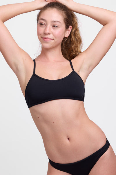 Female model wearing the Women’s Jocelyn Bikini Top in the Black solid, standing in front of a grey background, shown from front view. 
 
