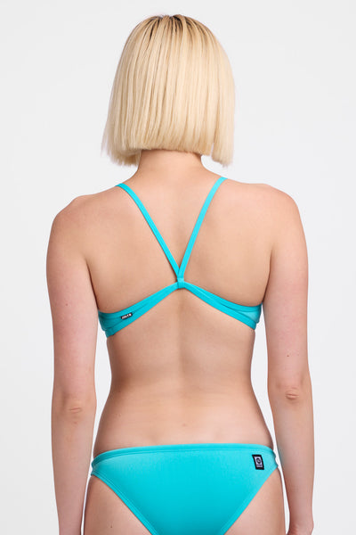 Female model wearing the Women’s Jocelyn Bikini Top in the Hawaii Blue solid, standing in front of a grey background, shown from back view. 
 