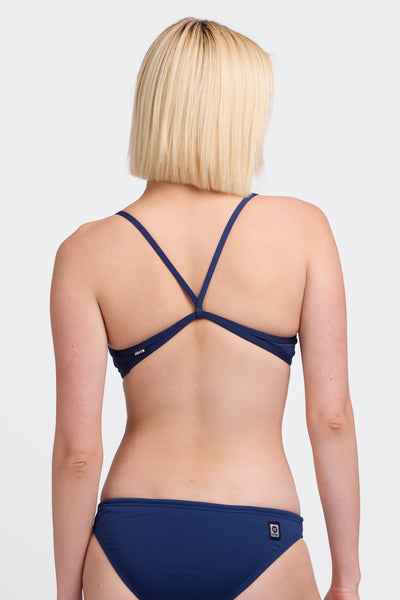 Female model wearing the Women’s Jocelyn Bikini Top in the Navy solid, standing in front of a grey background, shown from back view. 
 