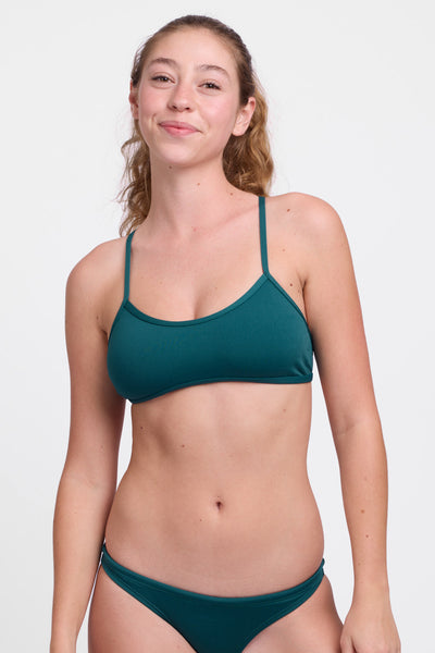 Female model wearing the Women’s Jocelyn Bikini Top in the Peacock solid, standing in front of a grey background, shown from front view. 
 