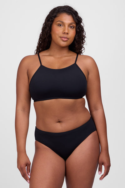 Female model wearing the Women’s Pam Bikini Bottoms in the Black solid, standing in front of a grey background, shown from front view. 
 