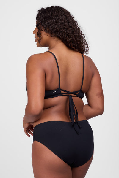 Female model wearing the Women’s Pam Bikini Bottoms in the Black solid, standing in front of a grey background, shown from back view. 
 