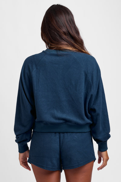 Female model wearing the Women's Maurie Crewneck in the Trade Winds solid, standing in front of a grey background, shown from the back view. 
