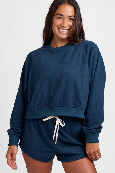 Female model wearing the Women's Maurie Crewneck in the Trade Winds solid, standing in front of a grey background, shown from the front view. 
