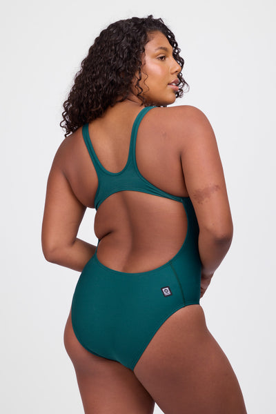 Female model wearing the Women’s Betty One Piece Swimsuit in the Peacock solid, standing in front of a grey background, shown from back view. 
