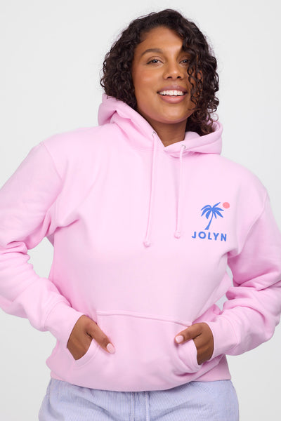 Female model wearing the Women’s Raine Hoodie in the Vibnin Pink solid, standing in front of a grey background, shown from front view. 
 