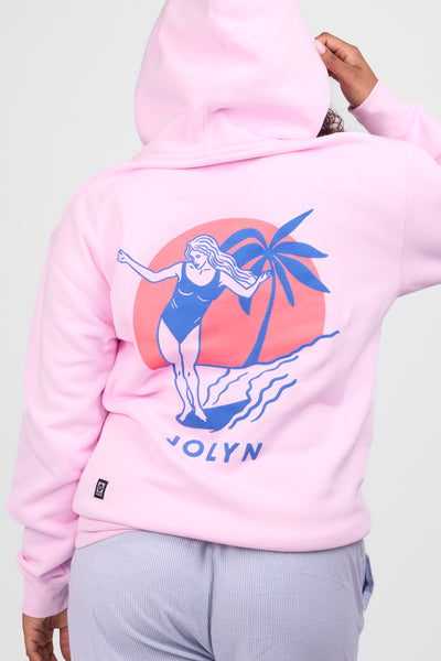 Female model wearing the Women’s Raine Hoodie in the Vibnin Pink solid, standing in front of a grey background, shown from back view. 
 