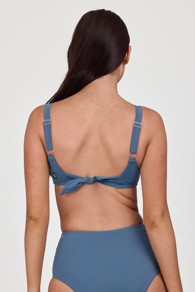 Female model wearing the Women’s Kacey Bikini Top in the Dolphins solid, standing in front of a grey background, shown from back view. 
 