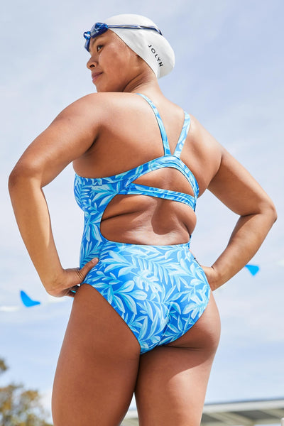 Female model wearing the Women's Caroline One Piece Swimsuit in the Canopy print, standing in front of a blue background, swim cap and goggles are on her head, shown from side view.