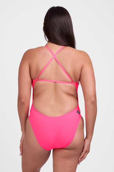 Female model wearing the Women's Brandon One Piece Swimsuit in the Hot Pink solid, standing in front of a grey background, shown from back view. 