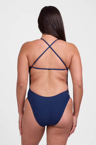Female model wearing the Women's Brandon One Piece Swimsuit in the Navy solid, standing in front of a grey background, shown from back view. 