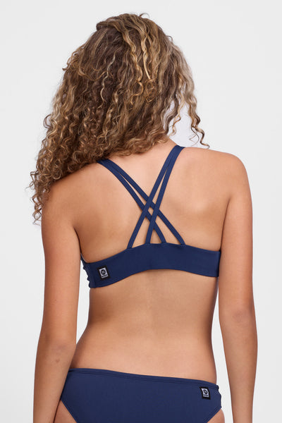 Female model wearing the Women’s Fendrick Bikini Top in the Navy solid, standing in front of a grey background, shown from back view. 