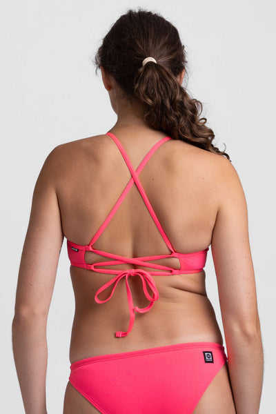 
Female model wearing the Women's Grayson Bikini Top in the Hot Pink solid, standing in front of a grey background, shown from back view. 
