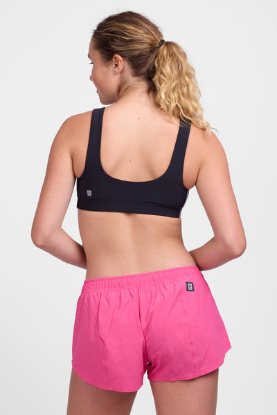 Female model wearing the Women’s Bennie Short in the Cerise solid, standing in front of a grey background, shown from back view. 