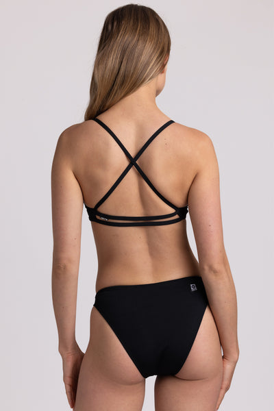 Female model wearing the Women’s Penny Bikini Bottom in the Black solid, standing in front of a grey background, shown from back view. 
