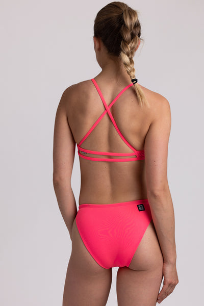 Female model wearing the Women’s Penny Bikini Bottom in the Hot Pink solid, standing in front of a grey background, shown from back view. 
