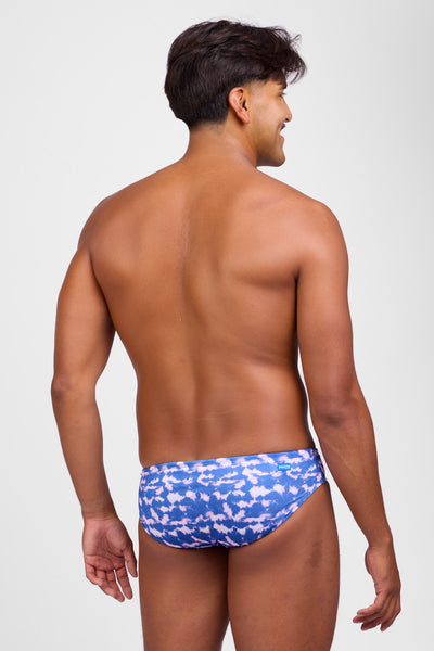 Female model wearing the Men's Brolyn Swim Brief in the Blue Tides print, standing in front of a grey background, shown from side view. 
