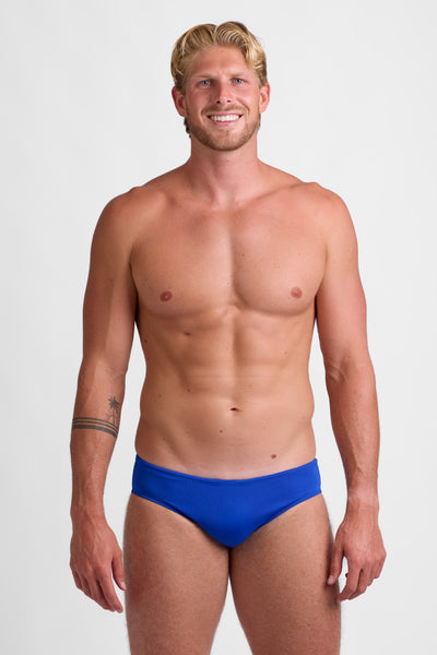 Male model wearing the Men's Brolyn Brief Swimsuit in the Blueberry solid, standing in front of a grey background, shown from front view. 