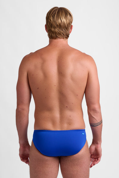 Male model wearing the Men's Brolyn Brief Swimsuit in the Blueberry solid, standing in front of a grey background, shown from back view. 
