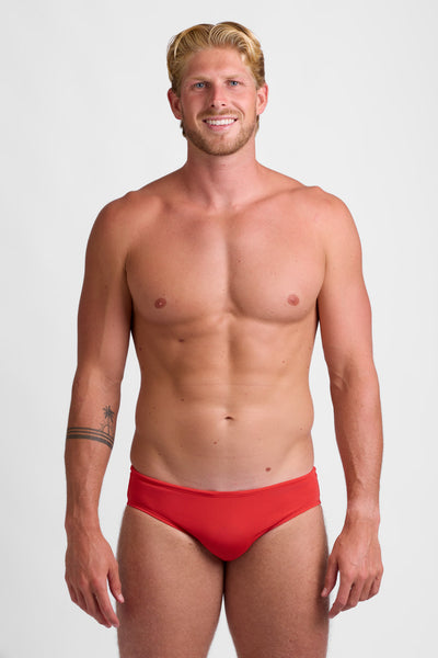 Male model wearing the Men's BROLYN Brief Swimsuit in the Candy Apple solid, standing in front of a grey background, shown from front view. 
