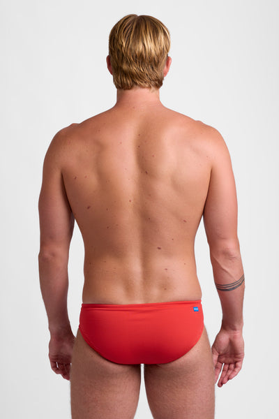 Male model wearing the Men's BROLYN Brief Swimsuit in the Candy Apple solid, standing in front of a grey background, shown from back view. 
