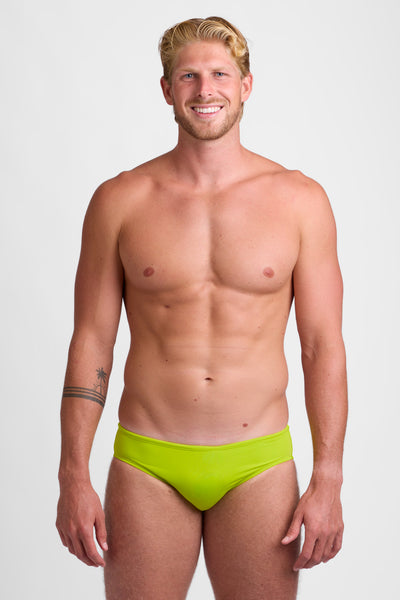Male model wearing the Men's BROLYN Brief Swimsuit in the Gecko solid, standing in front of a grey background, shown from front view. 
