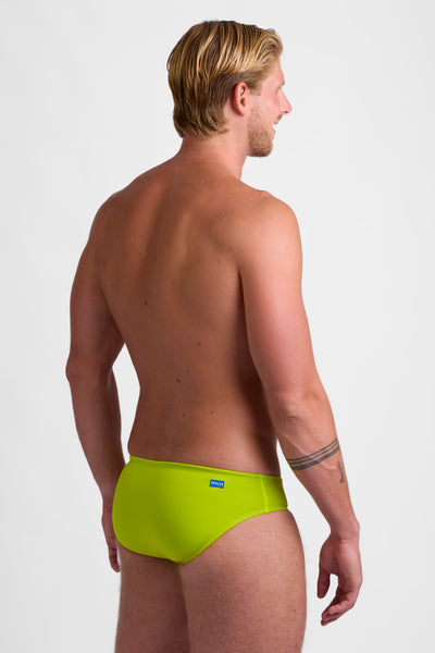 Male model wearing the Men's BROLYN Brief Swimsuit in the Gecko solid, standing in front of a grey background, shown from side view. 
