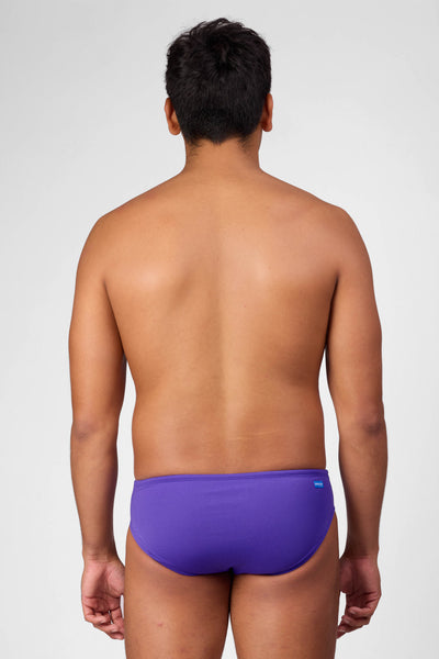Male model wearing the Men's Brolyn Swim Brief in the Purple solid, standing in front of a grey background shown from the back view