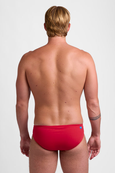 Male model wearing the Men's Brolyn Swim Brief in the Red solid, standing in front of a grey background shown from the back view