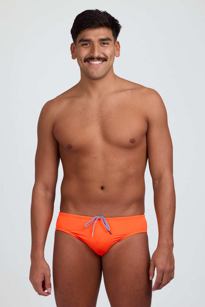 
Male model wearing the Men's Brolyn Swim Brief Swimsuit in the Sunkist solid, standing in front of a grey background shown from front view. 