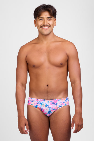Female model wearing the Men's Brolyn Swim Brief in the Superbloom print, standing in front of a grey background, shown from front view. 
