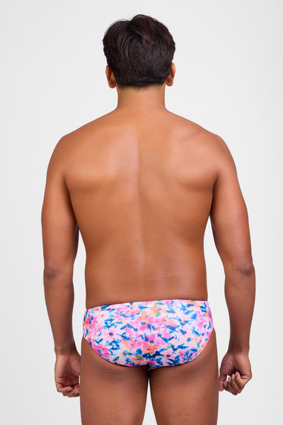 Female model wearing the Men's Brolyn Swim Brief in the Superbloom print, standing in front of a grey background, shown from back view. 

