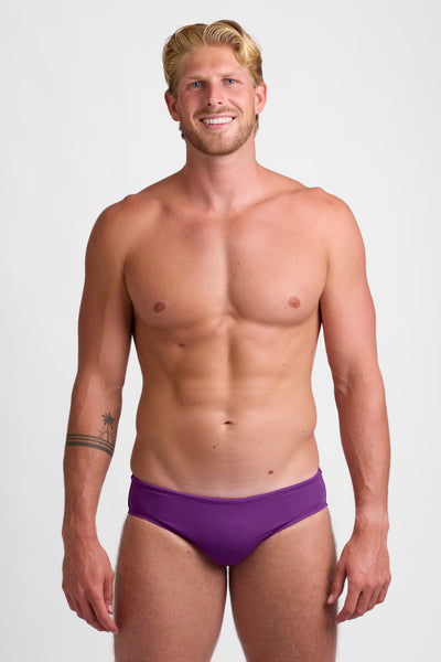 Male model wearing the Men's BROLYN Brief Swimsuit in the Verbena solid, standing in front of a grey background, shown from front view. 

