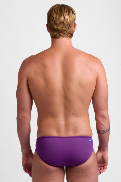 Male model wearing the Men's BROLYN Brief Swimsuit in the Verbena solid, standing in front of a grey background, shown from back view. 
