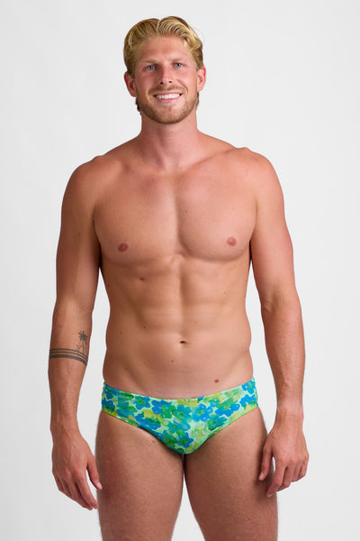 Male model wearing the Men's BROLYN Brief Swimsuit in the Water Blossom print, standing in front of a grey background, shown from front view. 