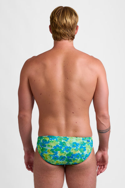 Male model wearing the Men's BROLYN Brief Swimsuit in the Water Blossom print, standing in front of a grey background, shown from back view. 