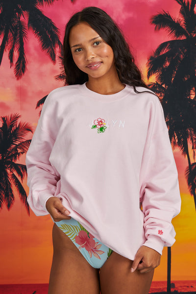 Female model wearing the Women’s Roxanne Crewneck in the Flower solid, standing in front of a sunset background, shown from front view. 