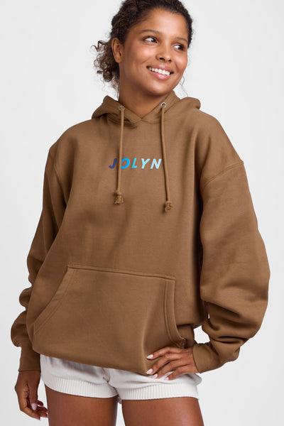 Female model wearing the Women's Raine Hoodie in the Toast Coco solid, standing in front of a grey background, shown from the front view. 