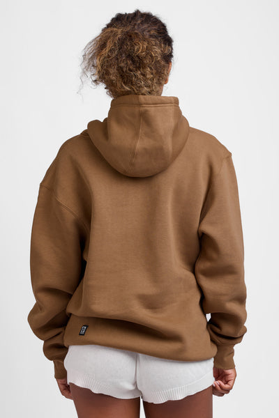 Female model wearing the Women's Raine Hoodie in the Toast Coco solid, standing in front of a grey background, shown from the back view. 