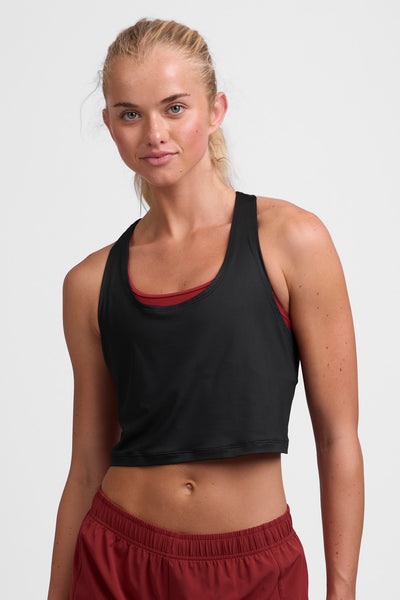 Female model wearing the Women’s Allison Run Crop Singlet in the Black solid, standing in front of a grey background, shown from front view. 