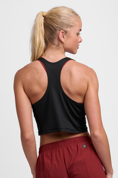 Female model wearing the Women’s Allison Run Crop Singlet in the Black solid, standing in front of a grey background, shown from back view. 