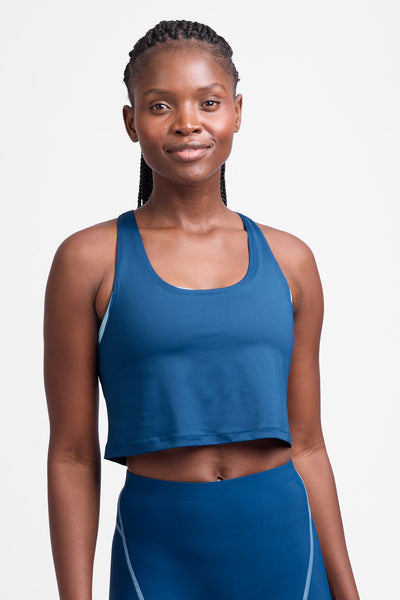 Female model wearing the Women’s Allison Run Crop Singlet in the Blue Flame solid, standing in front of a grey background, shown from front view. 