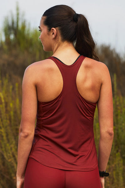 Female model wearing the Women’s Monti Run Singlet in the Spice solid, standing in front of a green background, shown from back view. 