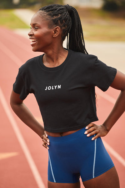 Female model wearing the Women's Delta T Shirt  in the JOLYN Logo print, standing in front of a track, shown from front view. 