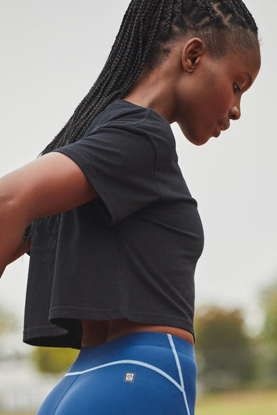 Female model wearing the Women's Delta T Shirt  in the JOLYN Logo print, standing in front of a track, shown from side view. 