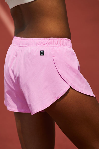 Female model wearing the Women's Lizzie Split Short in the Pinky solid, standing in front of a track, shown from the side view.